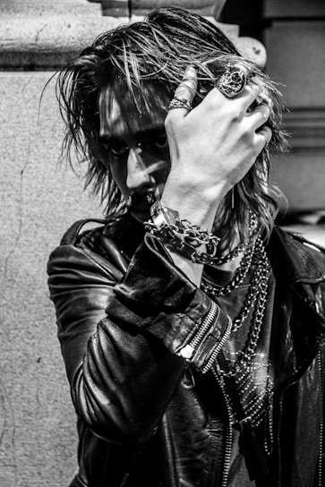 A person with an intense gaze is partially obscured by their hand, adorned with rings and bracelets. The individual wears a leather jacket and has long, textured hair, giving off a rock or punk aesthetic. Chains and jewelry add to the edgy look, and the photograph's black-and-white filter accentuates the mood.