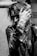 A person with an intense gaze is partially obscured by their hand, adorned with rings and bracelets. The individual wears a leather jacket and has long, textured hair, giving off a rock or punk aesthetic. Chains and jewelry add to the edgy look, and the photograph's black-and-white filter accentuates the mood.