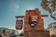 A stone monument stands with signage for Zion National Park, featuring a wooden sign with text and a National Park Service emblem with mountains and a bison. Surrounding the monument are trees and a clear blue sky, with distant mountain scenery in the background.