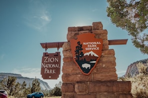 A stone monument stands with signage for Zion National Park, featuring a wooden sign with text and a National Park Service emblem with mountains and a bison. Surrounding the monument are trees and a clear blue sky, with distant mountain scenery in the background.