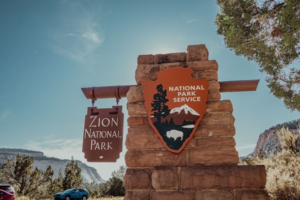 A stone monument stands with signage for Zion National Park, featuring a wooden sign with text and a National Park Service emblem with mountains and a bison. Surrounding the monument are trees and a clear blue sky, with distant mountain scenery in the background.