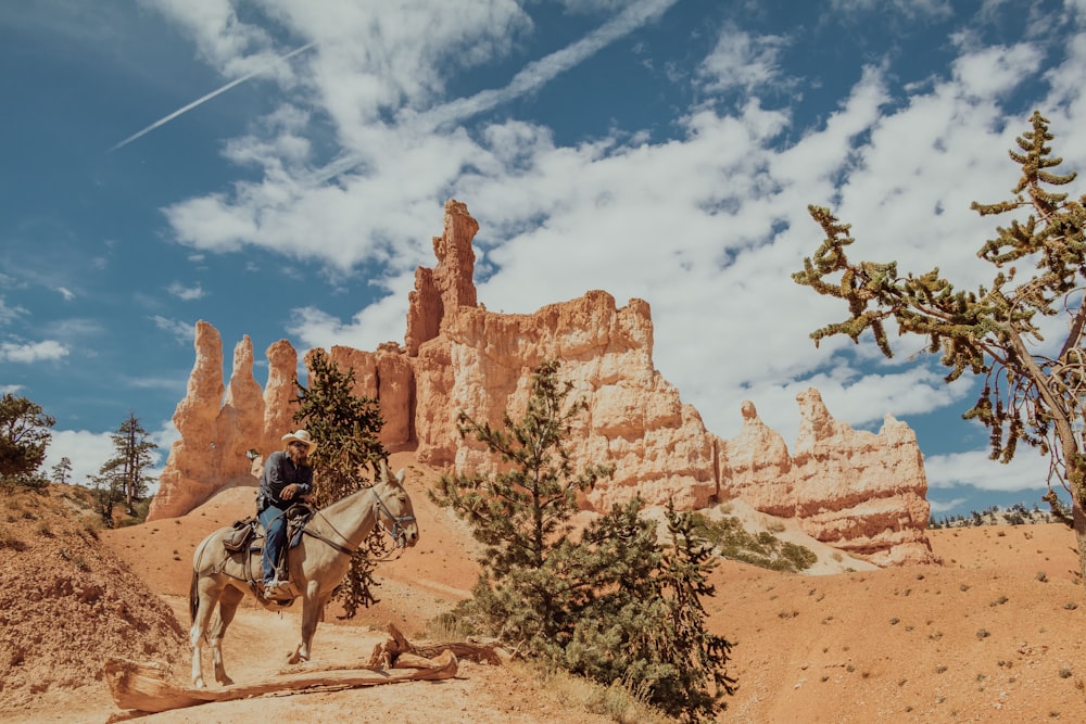 homem andando a cavalo na formação rochosa marrom sob o céu azul e nuvens brancas durante o dia