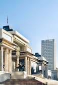 A large, modern building with classical architectural elements such as columns and statues of figures on horseback. The facade features reflective glass and golden accents. To the right, a tall, rectangular skyscraper rises against a clear blue sky. A flag flies atop the modern structure, adding a touch of national symbolism.