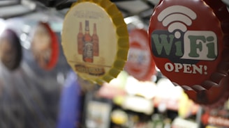 Several bottle cap-shaped signs are hanging from a ceiling. The most prominent sign is red and says 'WiFi OPEN!' with a WiFi symbol. Another visible sign has images of beer bottles on it.