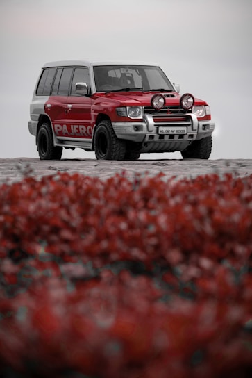 A red and white SUV is parked on a sandy surface, featuring off-road tires and additional front lights. In the foreground, there is a blurred patch of red vegetation or foliage.