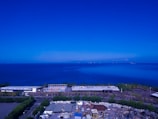 A vast body of water stretches out under a predominantly clear blue sky, with a few scattered clouds visible on the horizon. In the foreground, there appears to be an industrial area with buildings, roads, and trees. The ocean is expansive and a darker blue compared to the lighter sky above.