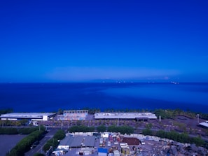 A vast body of water stretches out under a predominantly clear blue sky, with a few scattered clouds visible on the horizon. In the foreground, there appears to be an industrial area with buildings, roads, and trees. The ocean is expansive and a darker blue compared to the lighter sky above.
