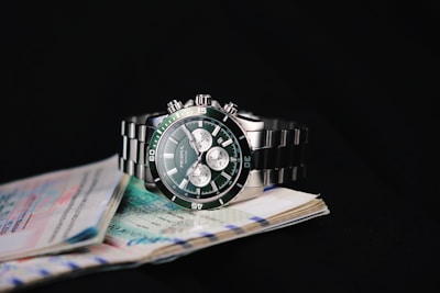 A sleek, metallic wristwatch rests on a stack of assorted documents against a dark background. The watch has a stainless steel band and a green dial with multiple sub-dials, indicating a sophisticated design.