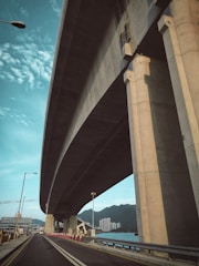 A large concrete highway overpass dominates the scene, curving gracefully above a stretch of roadway. Tall supporting pillars elevate the structure, giving it a sense of grandeur. Adjacent is a two-lane road lined with orange traffic cones. In the background are blue skies dotted with clouds and a distant view of city buildings near the water, with mountains rising on the horizon.