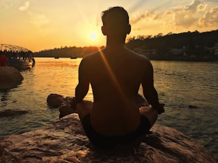 A person is meditating on a rock by the water at sunset, with the sun casting a golden glow across the scene. In the background, there are silhouettes of trees and a boat on the water, with clouds illuminated by the setting sun.