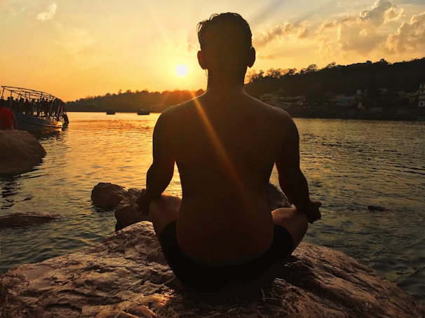 A person is meditating on a rock by the water at sunset, with the sun casting a golden glow across the scene. In the background, there are silhouettes of trees and a boat on the water, with clouds illuminated by the setting sun.