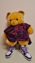 A plush teddy bear is dressed in a vibrant purple and pink shirt featuring a nature-inspired pattern. The bear is also wearing a pair of stylish high-top sneakers that are white and dark blue with purple laces.