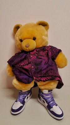 A plush teddy bear is dressed in a vibrant purple and pink shirt featuring a nature-inspired pattern. The bear is also wearing a pair of stylish high-top sneakers that are white and dark blue with purple laces.