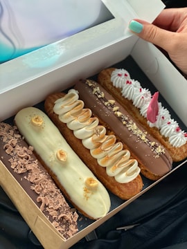 A box containing four assorted éclairs with various toppings is being held by a hand with teal nail polish. The éclairs are arranged in a row, each with unique toppings such as chocolate shavings, creamy swirls, chopped nuts, and decorative icing.