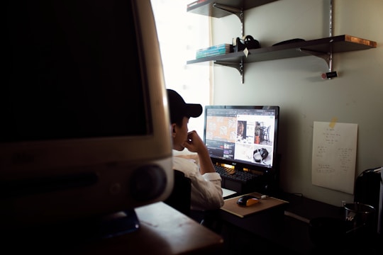A person wearing a cap is sitting at a desk, focusing on a computer monitor displaying various images. The workspace includes shelves with books and other items, a wall-mounted chart with notes, and another large monitor in the foreground.