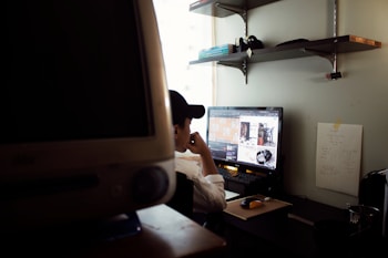 A person wearing a cap is sitting at a desk, focusing on a computer monitor displaying various images. The workspace includes shelves with books and other items, a wall-mounted chart with notes, and another large monitor in the foreground.