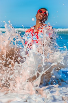 A person wearing a vibrant floral shirt and red sunglasses enjoys the ocean waves splashing around them. The water is captured in mid-splash, creating a dynamic and lively scene against a clear blue sky.