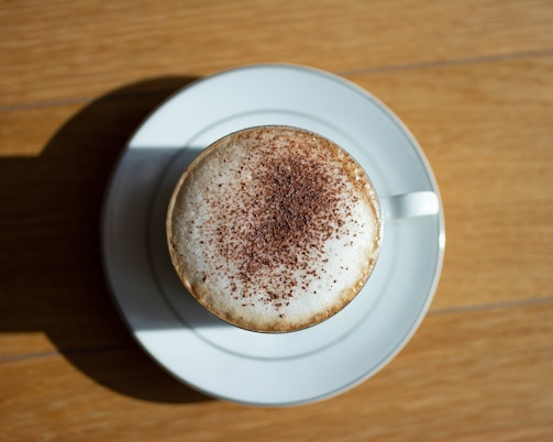 A cup of cappuccino with frothy milk topped with cocoa powder sits on a white saucer placed on a wooden table.