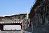 A large bridge under construction with extensive scaffolding and a highway passing underneath. Several vehicles are visible on the road. There are warning signs displaying a speed limit of 60. The sky is clear, and a banner displaying safety information is attached to the structure.