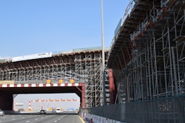 A large bridge under construction with extensive scaffolding and a highway passing underneath. Several vehicles are visible on the road. There are warning signs displaying a speed limit of 60. The sky is clear, and a banner displaying safety information is attached to the structure.