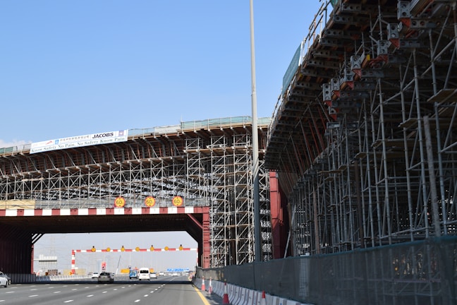 A large bridge under construction with extensive scaffolding and a highway passing underneath. Several vehicles are visible on the road. There are warning signs displaying a speed limit of 60. The sky is clear, and a banner displaying safety information is attached to the structure.