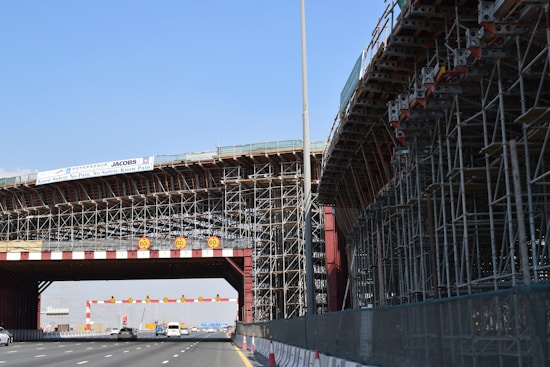 A large bridge under construction with extensive scaffolding and a highway passing underneath. Several vehicles are visible on the road. There are warning signs displaying a speed limit of 60. The sky is clear, and a banner displaying safety information is attached to the structure.