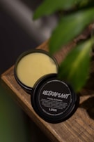 A container of facial cleanser labeled 'Ultraplant' from Lush sits open on a wooden surface, with a green plant partially visible in the background. The cleanser has a creamy, beige color, and the container is black with white text.