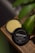 A container of facial cleanser labeled 'Ultraplant' from Lush sits open on a wooden surface, with a green plant partially visible in the background. The cleanser has a creamy, beige color, and the container is black with white text.