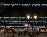 Night view of a lively plaza with string lights and people enjoying live music.