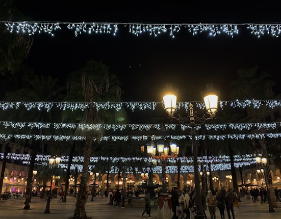A festive plaza illuminated by strings of white lights hanging overhead on a dark night. Tall lampposts with warm yellow lights provide additional illumination. Palm trees line the plaza, and groups of people are casually walking and gathering in the open space. Buildings in the background feature lit arched windows and bustling outdoor cafes.