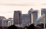 A cityscape featuring a cluster of modern skyscrapers with recognizable brand logos such as Citi, HSBC, and Barclays. The buildings have a mix of geometric shapes and glass facades, set against a backdrop of a cloudy sky during what appears to be dusk or dawn.