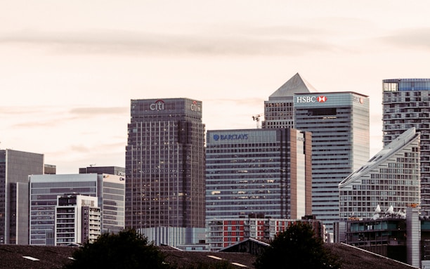 A cityscape featuring a cluster of modern skyscrapers with recognizable brand logos such as Citi, HSBC, and Barclays. The buildings have a mix of geometric shapes and glass facades, set against a backdrop of a cloudy sky during what appears to be dusk or dawn.