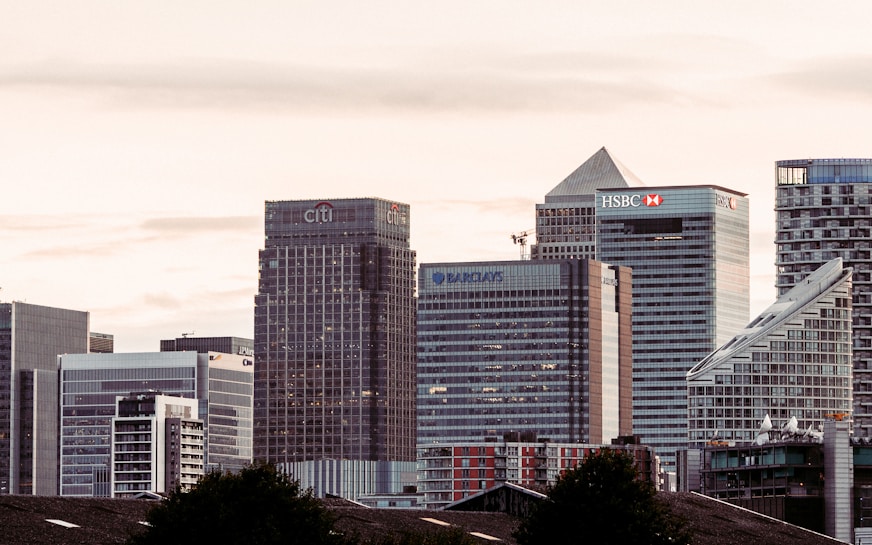 A cityscape featuring a cluster of modern skyscrapers with recognizable brand logos such as Citi, HSBC, and Barclays. The buildings have a mix of geometric shapes and glass facades, set against a backdrop of a cloudy sky during what appears to be dusk or dawn.