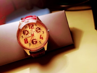 A wristwatch with a large, round face featuring bold Arabic numerals and a red leather strap is positioned on a rectangular, dark gray box. The face of the watch is cream-colored with red markings and a silver bezel, creating a vivid contrast. The background appears soft and out of focus, with warm lighting casting shadows that add depth to the image.