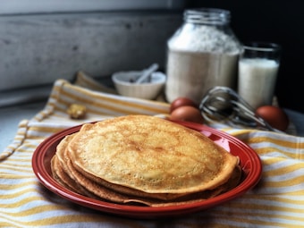 A stack of golden-brown pancakes is displayed on a red plate placed on a striped yellow and white cloth. In the background, there are baking ingredients including a jar of flour, a glass of milk, a whisk, eggs, and a small bowl with a spoon.
