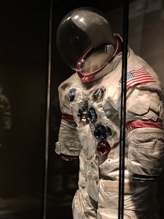 An astronaut suit displayed in a dimly lit museum setting. The suit is white with red and blue accents, featuring various patches including the NASA logo and an American flag on the shoulder. The helmet has a large, reflective visor, and the suit exhibits signs of wear, giving it a historical and iconic appearance.