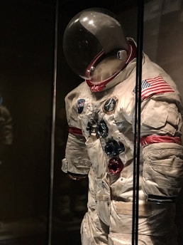 An astronaut suit displayed in a dimly lit museum setting. The suit is white with red and blue accents, featuring various patches including the NASA logo and an American flag on the shoulder. The helmet has a large, reflective visor, and the suit exhibits signs of wear, giving it a historical and iconic appearance.