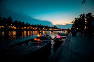 Rowboats are lined up along a calm river at dusk, with colorful lanterns glowing softly. The sky is transitioning from dark blue to lighter shades as evening falls. Trees and buildings along the waterfront are silhouetted against the twilight, and lights from the shore reflect on the water.