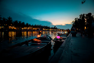 Rowboats are lined up along a calm river at dusk, with colorful lanterns glowing softly. The sky is transitioning from dark blue to lighter shades as evening falls. Trees and buildings along the waterfront are silhouetted against the twilight, and lights from the shore reflect on the water.
