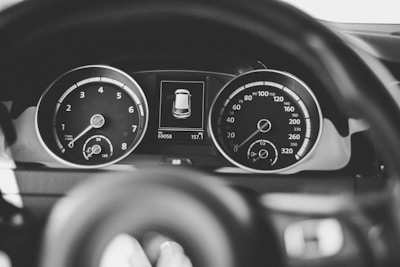 A close-up view of a car's dashboard featuring two large dials, one displaying the speedometer and the other the tachometer. An electronic display in the center shows an image of the car with mileage and other indicators.