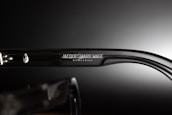 A close-up view of the side of a pair of eyeglasses showcasing the brand name 'Jacques Marie Mage' engraved on the temple along with a decorative emblem. The background is a smooth gradient from dark to light, emphasizing the glossy finish of the glasses.