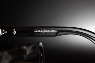 A close-up view of the side of a pair of eyeglasses showcasing the brand name 'Jacques Marie Mage' engraved on the temple along with a decorative emblem. The background is a smooth gradient from dark to light, emphasizing the glossy finish of the glasses.
