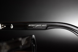 A close-up view of the side of a pair of eyeglasses showcasing the brand name 'Jacques Marie Mage' engraved on the temple along with a decorative emblem. The background is a smooth gradient from dark to light, emphasizing the glossy finish of the glasses.