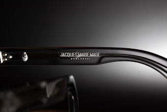 A close-up view of the side of a pair of eyeglasses showcasing the brand name 'Jacques Marie Mage' engraved on the temple along with a decorative emblem. The background is a smooth gradient from dark to light, emphasizing the glossy finish of the glasses.