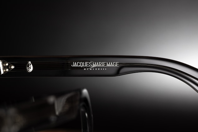 A close-up view of the side of a pair of eyeglasses showcasing the brand name 'Jacques Marie Mage' engraved on the temple along with a decorative emblem. The background is a smooth gradient from dark to light, emphasizing the glossy finish of the glasses.