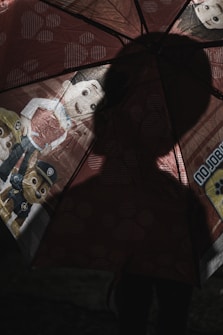 A shadowed figure stands behind a children's umbrella featuring cartoon characters in colorful attire. The lighting creates a silhouette effect, obscuring the identity of the person holding the umbrella.