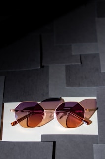 A pair of stylish sunglasses with pink and brown-tinted lenses rests on a surface composed of overlapping square paper tiles. The glasses have a rose-gold frame, and the light casts soft shadows, adding depth to the scene.