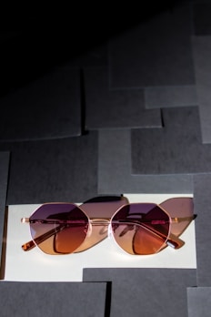 A pair of stylish sunglasses with pink and brown-tinted lenses rests on a surface composed of overlapping square paper tiles. The glasses have a rose-gold frame, and the light casts soft shadows, adding depth to the scene.