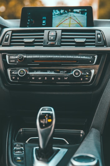 A car dashboard featuring a central control panel with various buttons, knobs, and a digital display. The display screen shows a rearview camera image with colored guide lines. Below the screen are controls for climate settings, radio, and media. The image also includes a gear shift lever with clearly visible markings and controls around it.