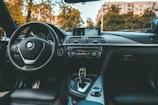 Interior of a BMW car showcasing the steering wheel, dashboard, multimedia controls, and gear shift. A navigation screen displays a rearview camera image. The image is taken from the driver's perspective, with trees and buildings visible through the windshield.